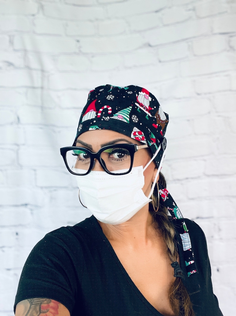 May include: A woman wearing a black scrub cap with a Christmas gnome pattern, black framed glasses, a white face mask, and a black shirt. Her hair is braided and tied with a matching Christmas gnome patterned fabric.