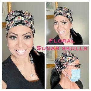 May include: A black scrub hat with a colorful floral pattern featuring sugar skulls. The hat has a tie at the back and a button on the side. The hat is worn by a person with long black hair. The text "FLORAL SUGAR SKULLS" is visible in the image.