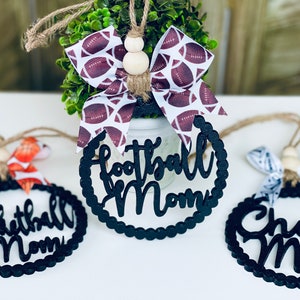 Puede incluir: Tres adornos de madera negra con borde festoneado y una cuerda blanca para colgar. Los adornos est&aacute;n decorados con las palabras "Football Mom", "Basketball Mom" y "Cheer Mom".