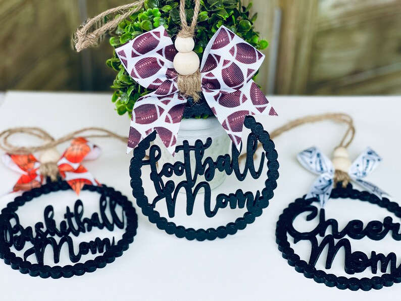 Puede incluir: Tres adornos de madera negra con borde festoneado y una cuerda blanca para colgar. Los adornos est&aacute;n decorados con las palabras "Football Mom", "Basketball Mom" y "Cheer Mom".