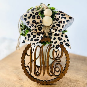 May include: A wooden ornament with a monogram in the center, surrounded by a beaded border. The ornament is decorated with a leopard print bow and two wooden beads.