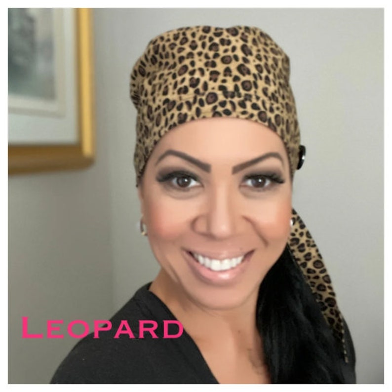 May include: A brown and black leopard print surgical scrub hat with a button on the side. The hat has a long tie that can be tied at the back of the neck.