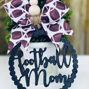 Puede incluir: Adorno de madera negro con las palabras "Football Mom" en una fuente decorativa. El adorno est&aacute; decorado con un lazo blanco y burdeos con un patr&oacute;n de f&uacute;tbol.