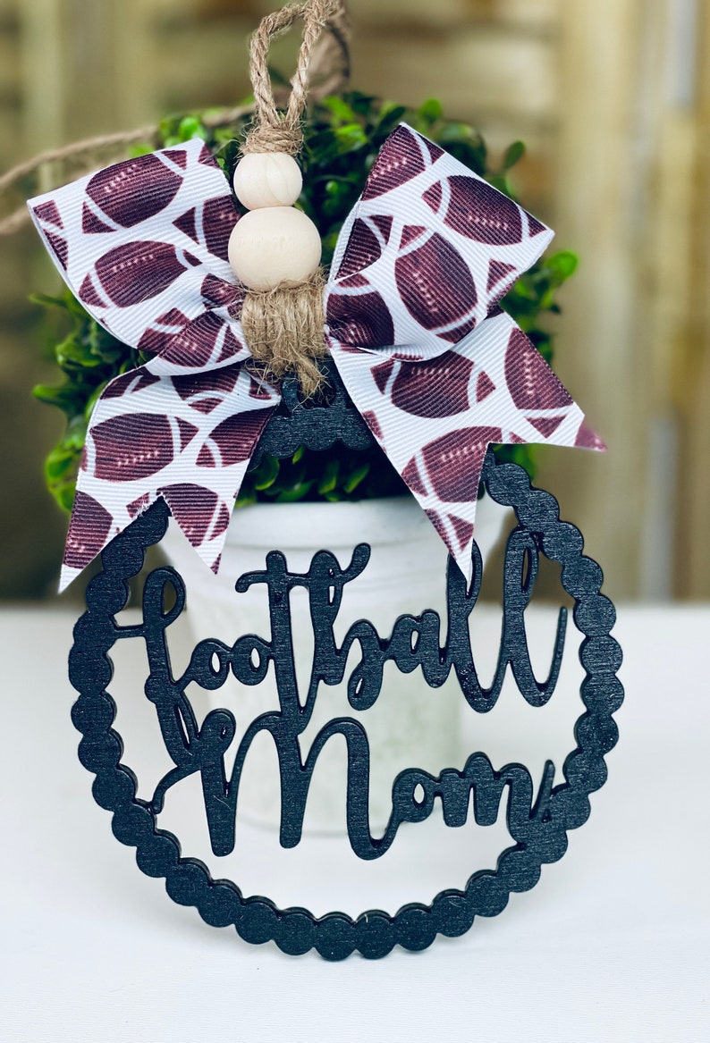 Puede incluir: Adorno de madera negro con las palabras "Football Mom" en una fuente decorativa. El adorno est&aacute; decorado con un lazo blanco y burdeos con un patr&oacute;n de f&uacute;tbol.