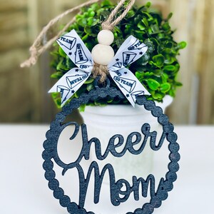 Puede incluir: Adorno de madera negro con las palabras "Cheer Mom" en una fuente decorativa. El adorno cuelga de una maceta blanca con una planta verde en su interior. El adorno est&aacute; decorado con una cinta blanca con texto negro que dice "Go Team".