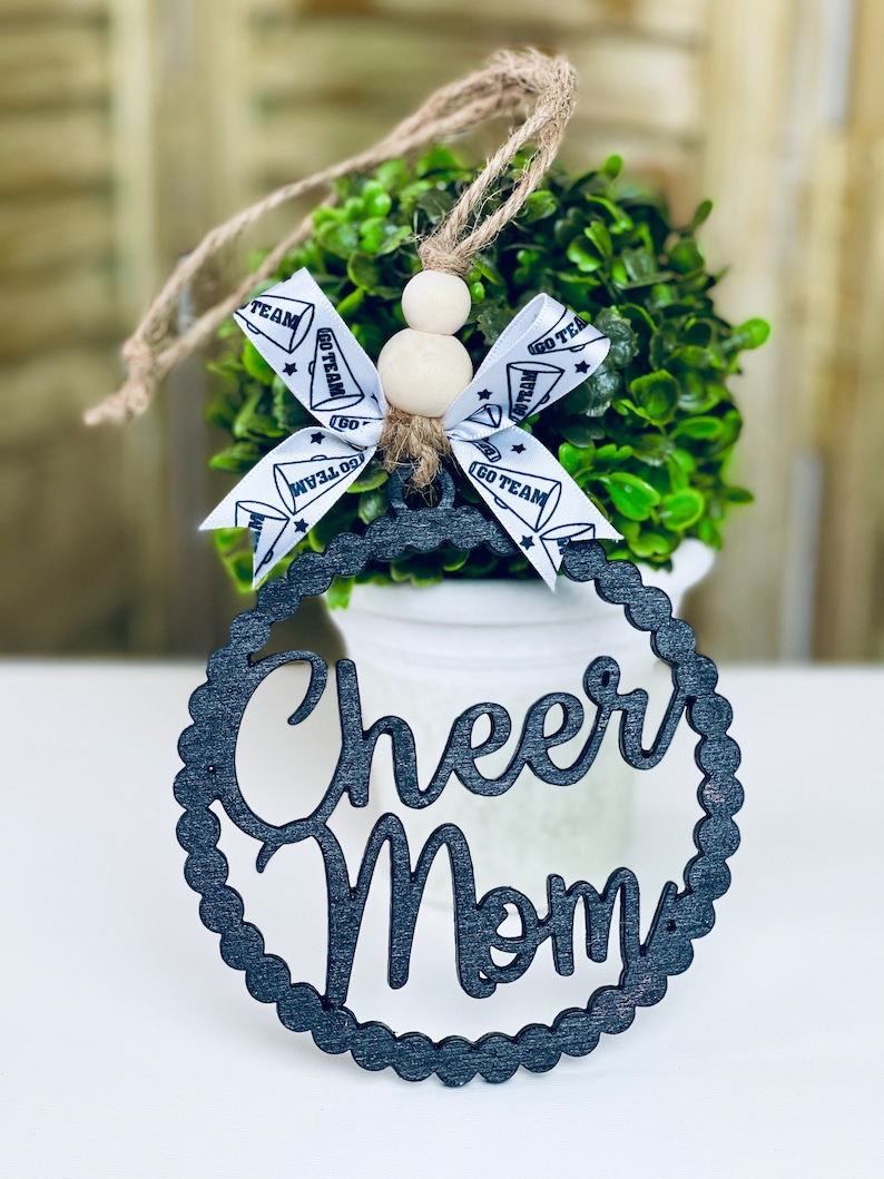 Puede incluir: Adorno de madera negro con las palabras "Cheer Mom" en una fuente decorativa. El adorno cuelga de una maceta blanca con una planta verde en su interior. El adorno est&aacute; decorado con una cinta blanca con texto negro que dice "Go Team".