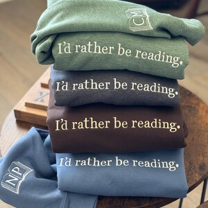 K&ouml;nnte beinhalten: Ein Stapel gefalteter Sweatshirts in Gr&uuml;n, Grau, Braun und Blau. Jedes Sweatshirt hat den gestickten Satz "I'd rather be reading." Die &Auml;rmel zeigen gestickte Buch- und Initialen-Designs. Die Sweatshirts sind auf einem Holztisch gestapelt.