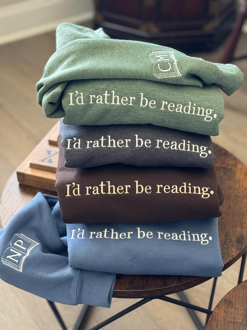 K&ouml;nnte beinhalten: Ein Stapel gefalteter Sweatshirts in Gr&uuml;n, Grau, Braun und Blau. Jedes Sweatshirt hat den gestickten Satz "I'd rather be reading." Die &Auml;rmel zeigen gestickte Buch- und Initialen-Designs. Die Sweatshirts sind auf einem Holztisch gestapelt.