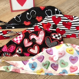 May include: Four Valentine's Day themed surgical scrub hats with different heart patterns. The hats are black with red hearts, black and white stripes with red hearts, black with pink and red hearts, and white with colorful candy hearts. Each hat has a button closure.