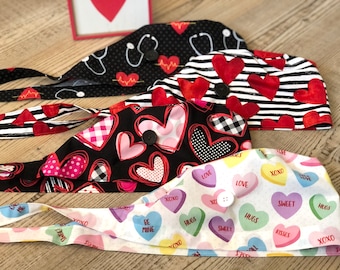 May include: Four Valentine's Day themed surgical scrub hats with different heart patterns. The hats are black with red hearts, black and white stripes with red hearts, black with pink and red hearts, and white with colorful candy hearts. Each hat has a button closure.
