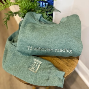 K&ouml;nnte beinhalten: Zwei gr&uuml;ne Sweatshirts werden gezeigt, eines gefaltet und das andere teilweise entfaltet. Das gefaltete Sweatshirt hat den gestickten Text "I'd rather be reading". Der &Auml;rmel des anderen Sweatshirts hat ein gesticktes Buchdesign mit den Buchstaben "MM".