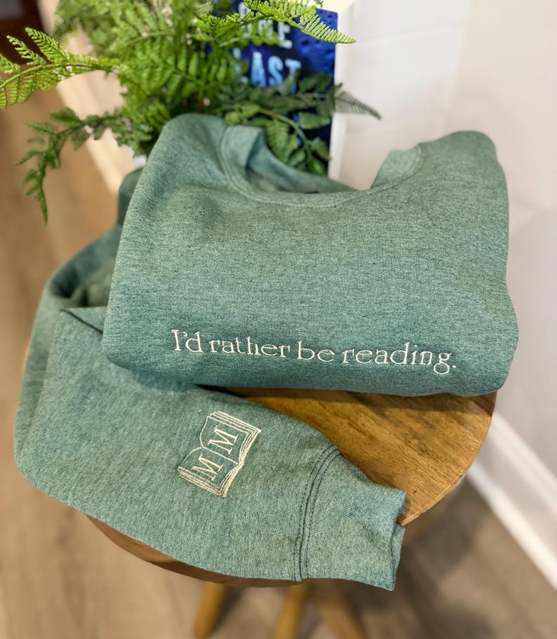 K&ouml;nnte beinhalten: Zwei gr&uuml;ne Sweatshirts werden gezeigt, eines gefaltet und das andere teilweise entfaltet. Das gefaltete Sweatshirt hat den gestickten Text "I'd rather be reading". Der &Auml;rmel des anderen Sweatshirts hat ein gesticktes Buchdesign mit den Buchstaben "MM".