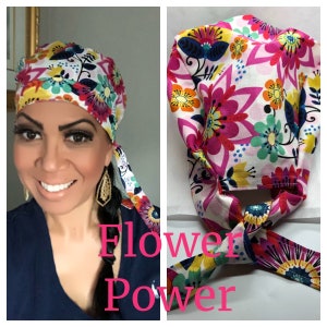 May include: A colorful floral print surgical cap with a tie. The cap is white with pink, yellow, blue, and green flowers. The tie is tied in a bow at the back of the neck. The text "Flower Power" is written in pink letters below the cap.