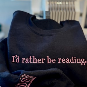 K&ouml;nnte beinhalten: Schwarzes Sweatshirt mit dem in Rosa gestickten Schriftzug "I'd rather be reading". Der &Auml;rmel zeigt ein rosa gesticktes Buch und die Initialen "DS". Das Sweatshirt ist gefaltet.