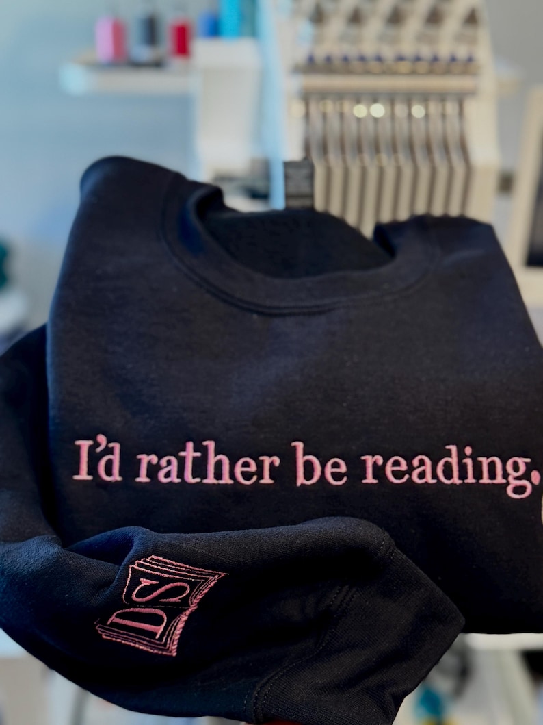 K&ouml;nnte beinhalten: Schwarzes Sweatshirt mit dem in Rosa gestickten Schriftzug "I'd rather be reading". Der &Auml;rmel zeigt ein rosa gesticktes Buch und die Initialen "DS". Das Sweatshirt ist gefaltet.