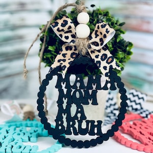 May include: Black wooden ornament with the words "Mama Mom Ma  Bruh" in a circle. The ornament is decorated with a leopard print bow and white beads.