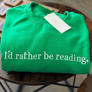 K&ouml;nnte beinhalten: Hellgr&uuml;nes Sweatshirt mit dem gestickten Text "I'd rather be reading." Ein wei&szlig;es Etikett ist am Sweatshirt befestigt. Das Sweatshirt liegt gefaltet auf einem dunkelbraunen Holztisch.