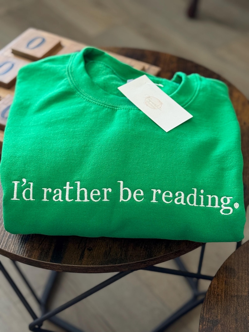 K&ouml;nnte beinhalten: Hellgr&uuml;nes Sweatshirt mit dem gestickten Text "I'd rather be reading." Ein wei&szlig;es Etikett ist am Sweatshirt befestigt. Das Sweatshirt liegt gefaltet auf einem dunkelbraunen Holztisch.