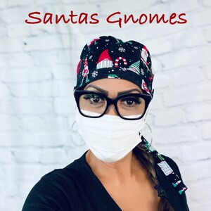 May include: A black scrub hat with a Christmas gnome pattern. The hat has a white background with red, green, and white gnomes. The hat is worn by a person wearing a black shirt and a white face mask. The text "Santas Gnomes" is written in red at the top of the image.