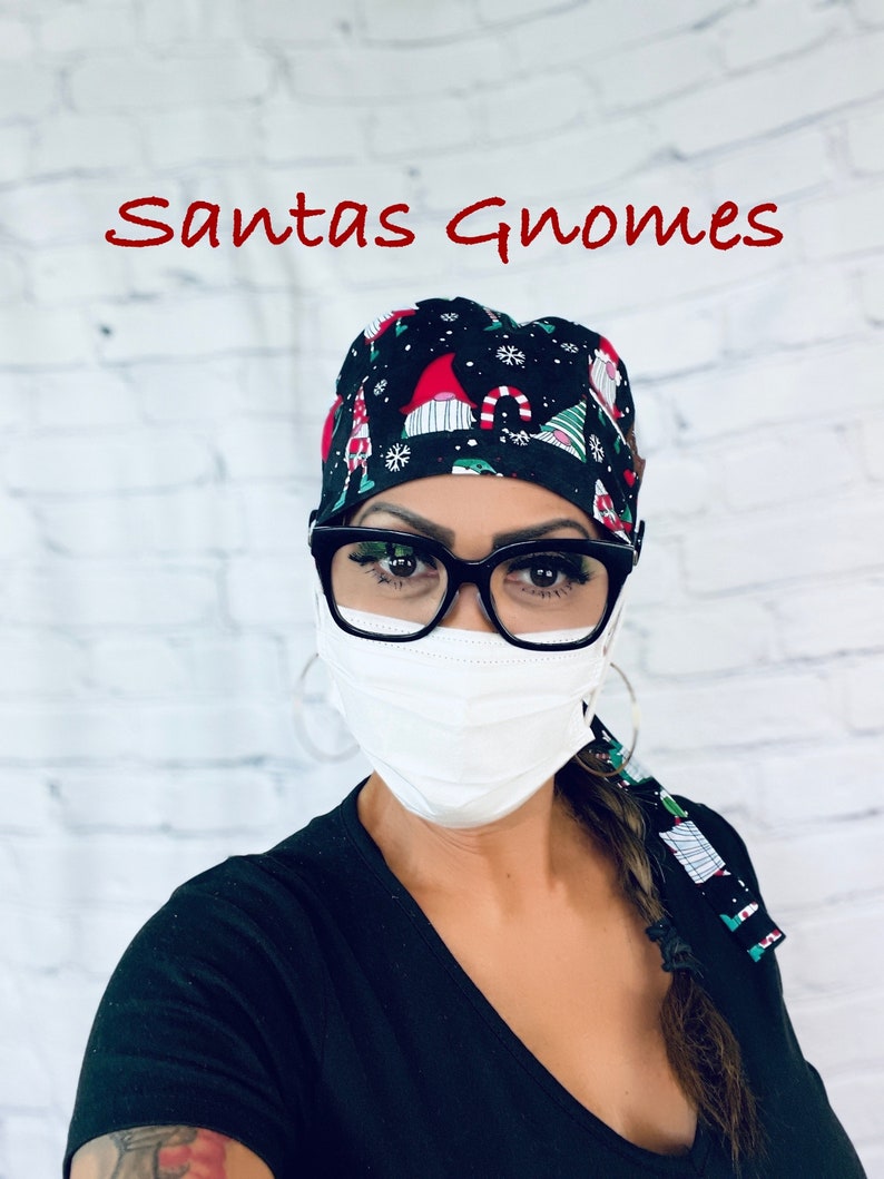 May include: A black scrub hat with a Christmas gnome pattern. The hat has a white background with red, green, and white gnomes. The hat is worn by a person wearing a black shirt and a white face mask. The text "Santas Gnomes" is written in red at the top of the image.