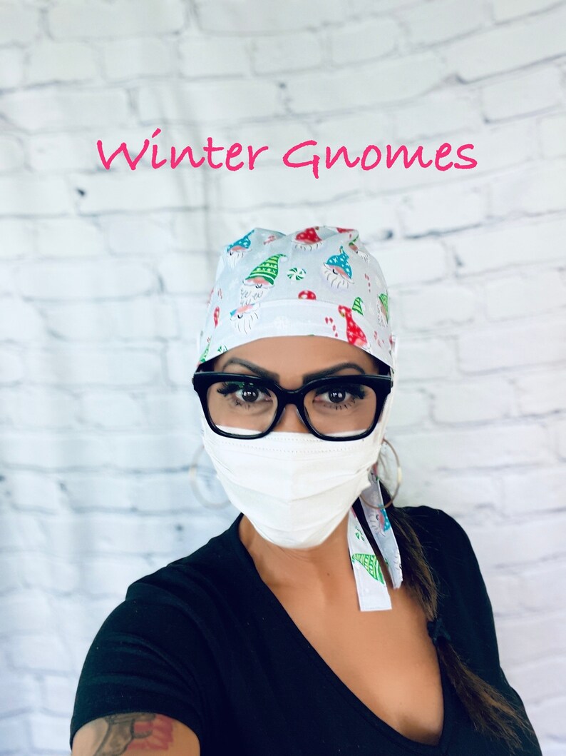 May include: A white scrub cap with a pattern of winter gnomes wearing red hats and white beards. The cap has a tie that is tied around the back of the head. The person wearing the cap is also wearing a white face mask and black shirt.