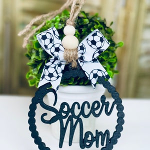 Puede incluir: Lazo de cinta con estampado de bal&oacute;n de f&uacute;tbol blanco y negro con cuentas de madera en un arbusto verde. Un letrero de madera negro con las palabras "Soccer Mom" en un c&iacute;rculo.