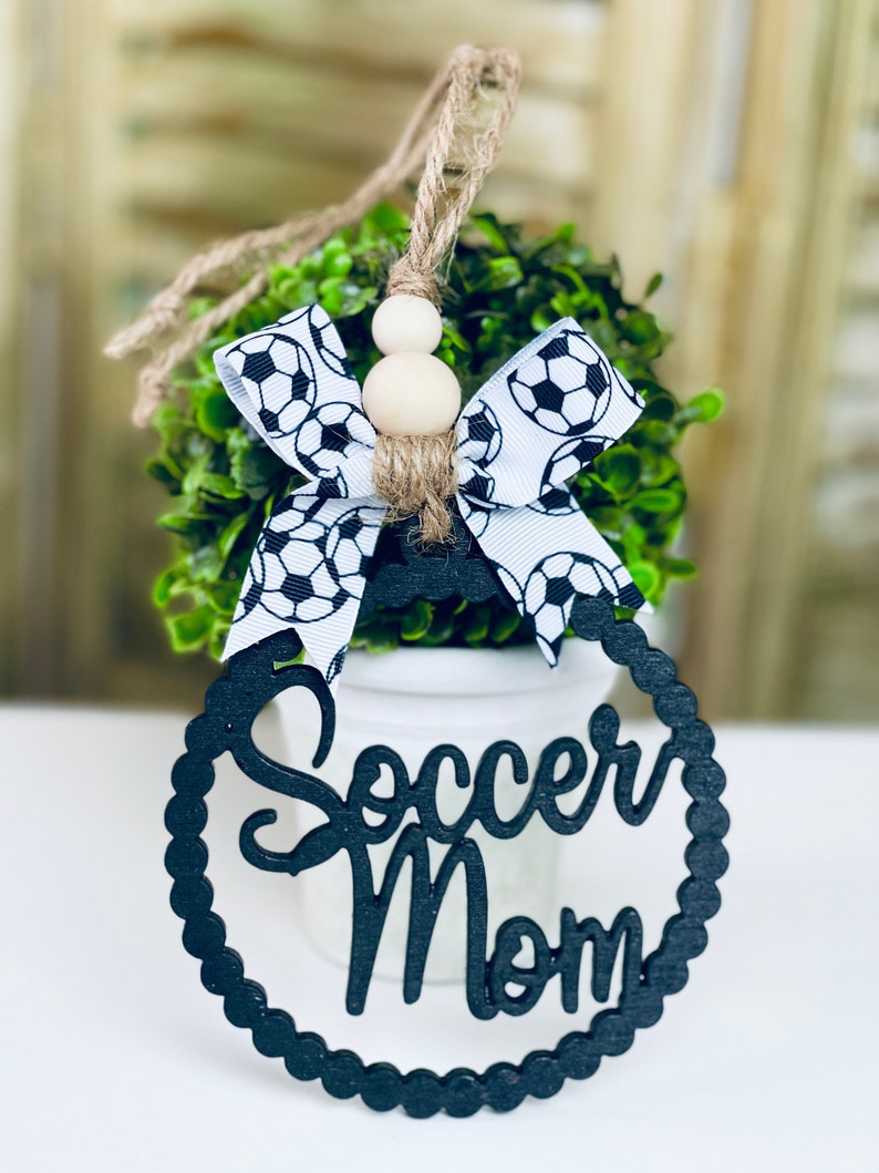 Puede incluir: Lazo de cinta con estampado de bal&oacute;n de f&uacute;tbol blanco y negro con cuentas de madera en un arbusto verde. Un letrero de madera negro con las palabras "Soccer Mom" en un c&iacute;rculo.