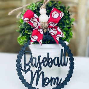 Puede incluir: Adorno de madera negro con las palabras "Baseball Mom" en una fuente decorativa. El adorno est&aacute; decorado con un lazo rojo y blanco con tem&aacute;tica de b&eacute;isbol.