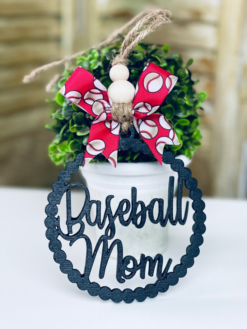 Puede incluir: Adorno de madera negro con las palabras "Baseball Mom" en una fuente decorativa. El adorno est&aacute; decorado con un lazo rojo y blanco con tem&aacute;tica de b&eacute;isbol.