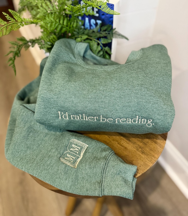 K&ouml;nnte beinhalten: Zwei gr&uuml;ne Sweatshirts werden gezeigt, eines gefaltet und das andere teilweise entfaltet. Das gefaltete Sweatshirt hat den gestickten Text "I'd rather be reading". Der &Auml;rmel des anderen Sweatshirts hat ein gesticktes Buchdesign mit den Buchstaben "MM".