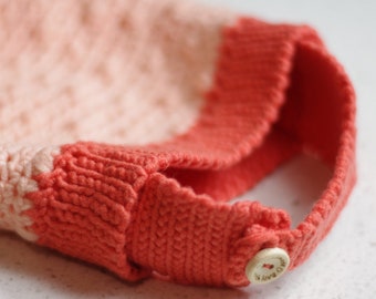 May include: A crocheted textile item featuring a light peach-colored main section and a vibrant coral-colored ribbed border. A small, round, light-colored wooden button is attached to the coral-colored strap, featuring the text "made with love".