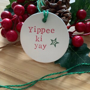 May include: A round, off-white Christmas ornament with the phrase "Yippee ki yay" in red letters and a green star. The ornament is strung with green twine and is surrounded by red berries, green leaves, and a pine cone.