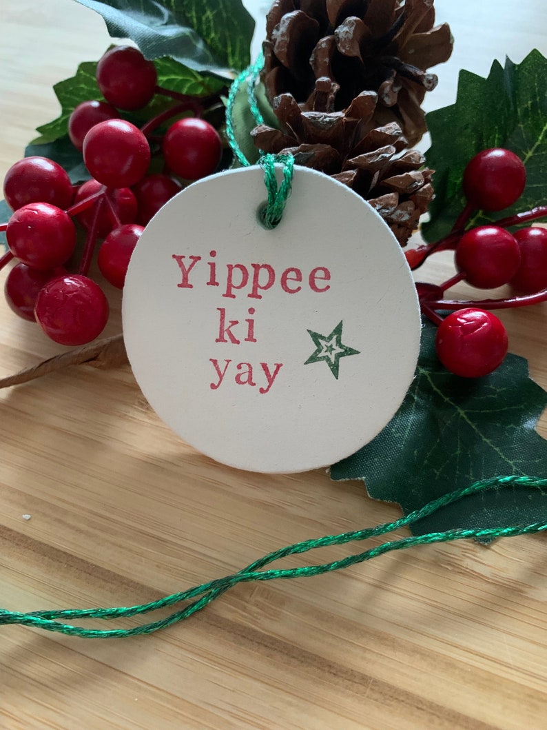 May include: A round, off-white Christmas ornament with the phrase "Yippee ki yay" in red letters and a green star. The ornament is strung with green twine and is surrounded by red berries, green leaves, and a pine cone.