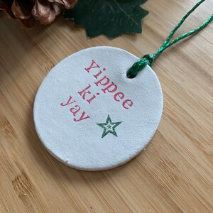 May include: A round, white Christmas ornament with the phrase "Yippee ki yay" in red lettering and a green star. The ornament has a green string for hanging. The background includes a pinecone, holly, and a wooden surface.
