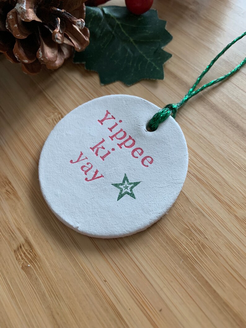 May include: A round, white Christmas ornament with the phrase "Yippee ki yay" in red lettering and a green star. The ornament has a green string for hanging. The background includes a pinecone, holly, and a wooden surface.