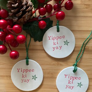 May include: Three white, round Christmas ornaments with the text "Yippee ki yay" in pink and a green star. The ornaments are hanging with green string. The background includes red berries, green leaves, and a pine cone.
