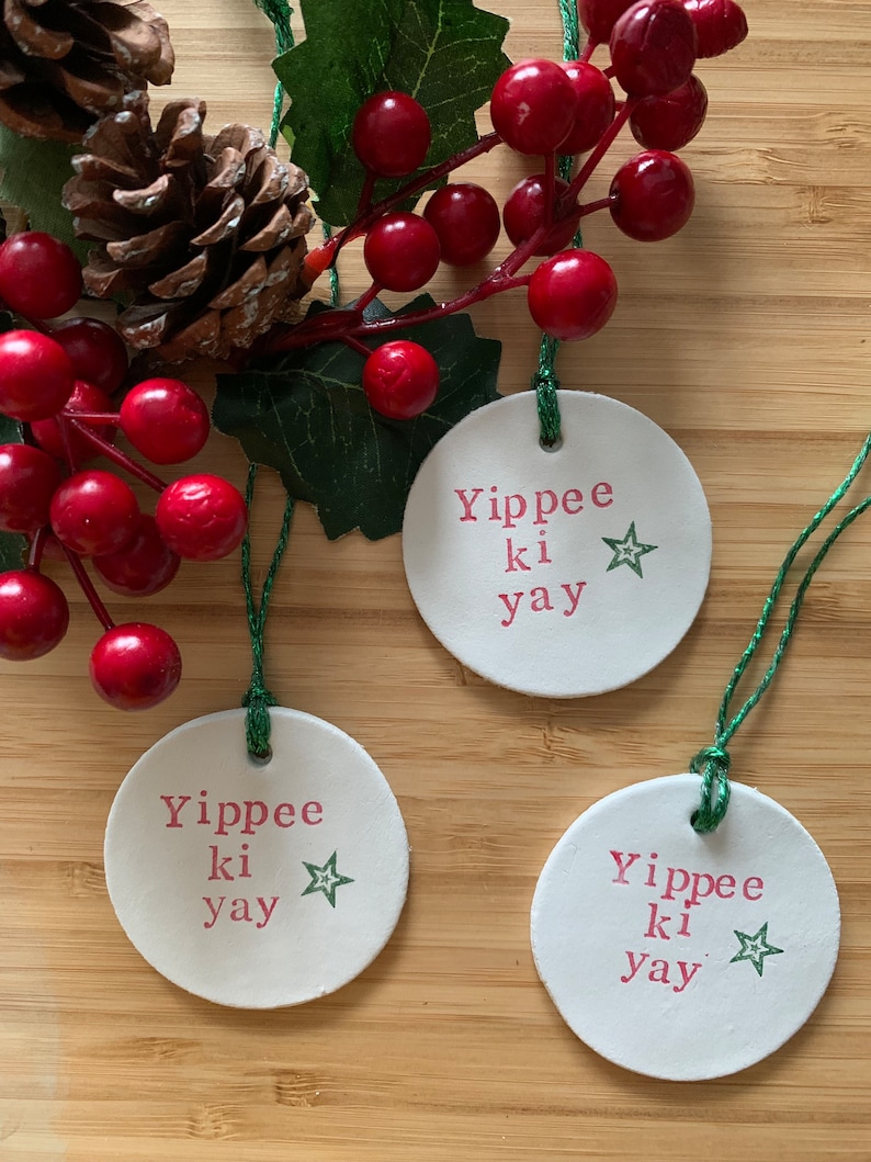 May include: Three white, round Christmas ornaments with the text "Yippee ki yay" in pink and a green star. The ornaments are hanging with green string. The background includes red berries, green leaves, and a pine cone.