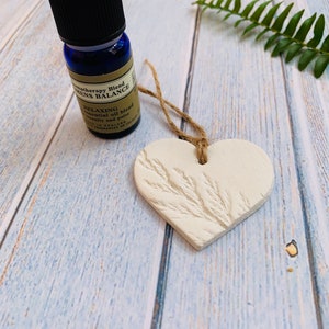 May include: A white ceramic heart-shaped ornament with a twig design, hanging from a twine string. A small blue glass bottle with a black cap and a label that reads "Aromatherapy Blend Womens Balance Relaxing Essential Oil Blend" is in the background.