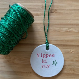 May include: A white, round ornament with the phrase "Yippee ki yay" in pink lettering and a green star. A spool of green glittery twine and a length of green twine are also visible.