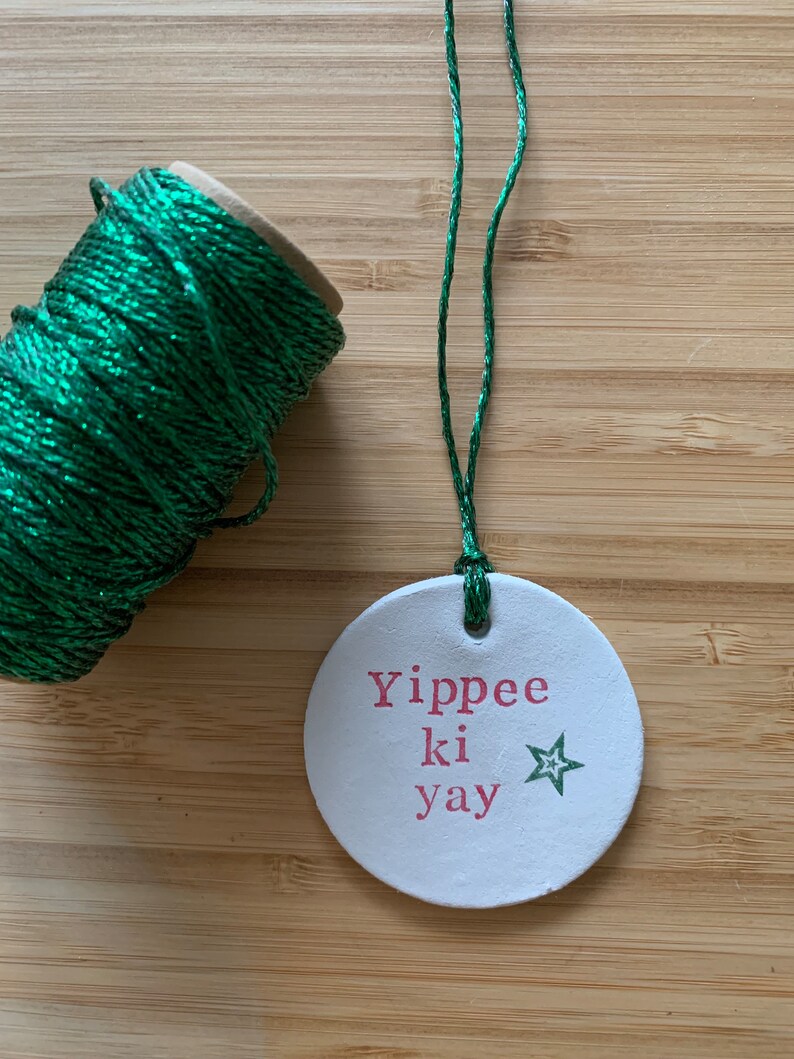 May include: A white, round ornament with the phrase "Yippee ki yay" in pink lettering and a green star. A spool of green glittery twine and a length of green twine are also visible.