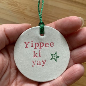 May include: A round, off-white Christmas ornament with the phrase "Yippee ki yay" in red lettering and a green star. The ornament is strung with a green cord.