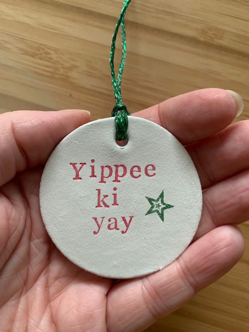 May include: A round, off-white Christmas ornament with the phrase "Yippee ki yay" in red lettering and a green star. The ornament is strung with a green cord.