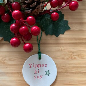 May include: A white, round Christmas ornament with the phrase "Yippee ki yay" in pink letters and a green star. The ornament hangs from a green string and is surrounded by red berries, green leaves, and a pine cone.