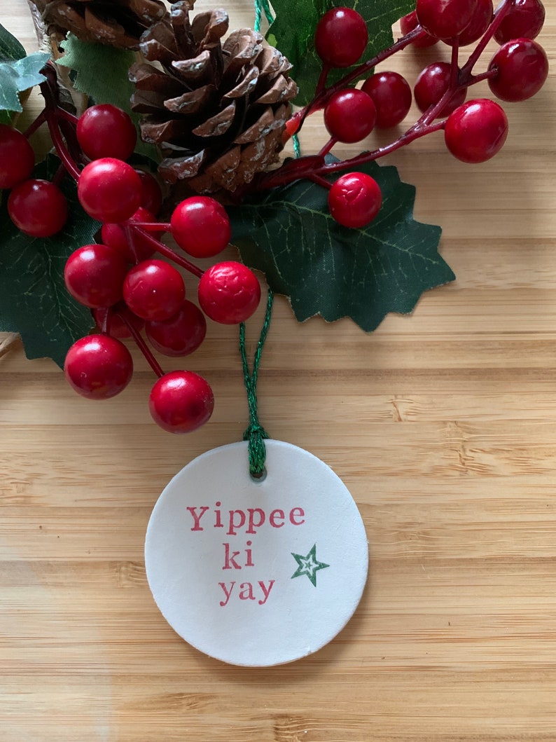 May include: A white, round Christmas ornament with the phrase "Yippee ki yay" in pink letters and a green star. The ornament hangs from a green string and is surrounded by red berries, green leaves, and a pine cone.
