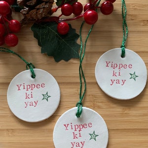 May include: Three white, round Christmas ornaments with the phrase "Yippee ki yay" in pink lettering and a green star. Each ornament has a green string for hanging. The ornaments are arranged on a wooden surface with red berries and a pinecone.