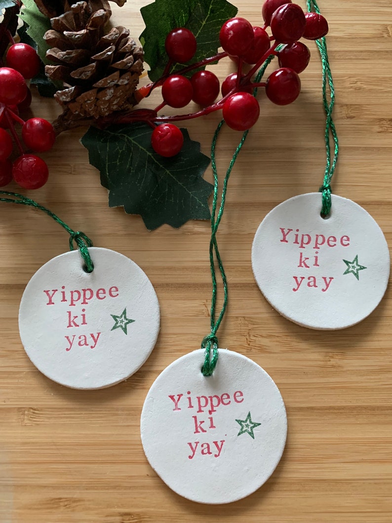May include: Three white, round Christmas ornaments with the phrase "Yippee ki yay" in pink lettering and a green star. Each ornament has a green string for hanging. The ornaments are arranged on a wooden surface with red berries and a pinecone.