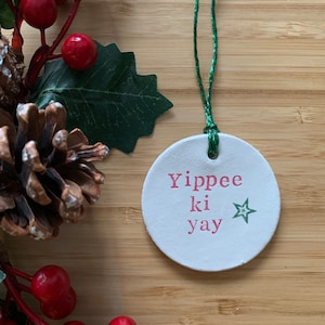 May include: A round, white Christmas ornament with the phrase "Yippee ki yay" in pink lettering and a green star. The ornament hangs from a green string and is surrounded by red berries, green leaves, and a pinecone.