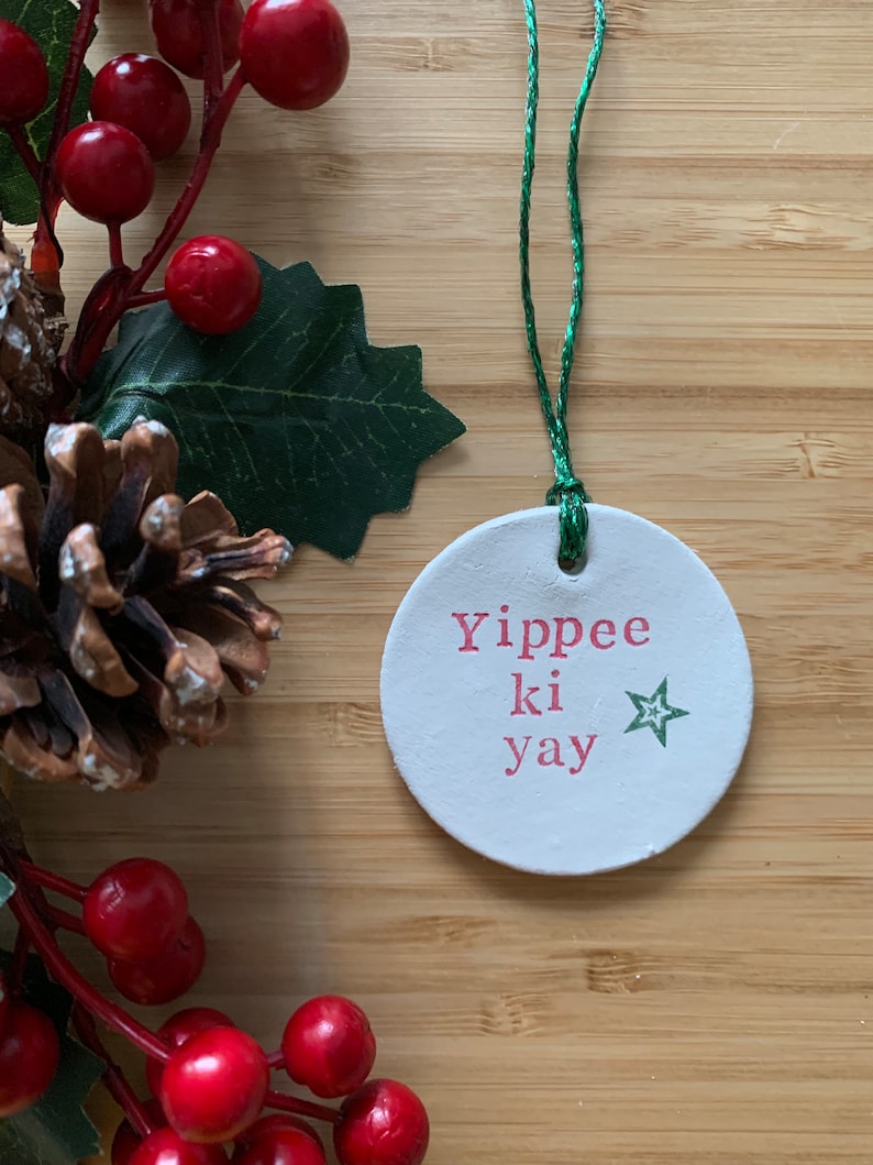 May include: A round, white Christmas ornament with the phrase "Yippee ki yay" in pink lettering and a green star. The ornament hangs from a green string and is surrounded by red berries, green leaves, and a pinecone.