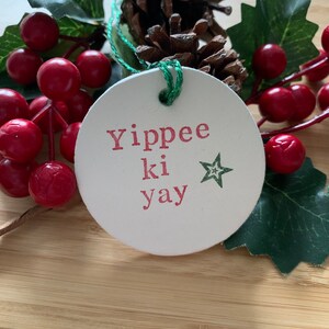 May include: A round, off-white Christmas ornament with the phrase "Yippee ki yay" in red letters and a green star. The ornament is attached with green string and is surrounded by red berries, green leaves, and a pinecone.