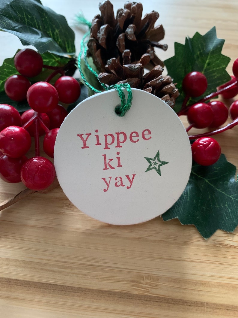 May include: A round, off-white Christmas ornament with the phrase "Yippee ki yay" in red letters and a green star. The ornament is attached with green string and is surrounded by red berries, green leaves, and a pinecone.